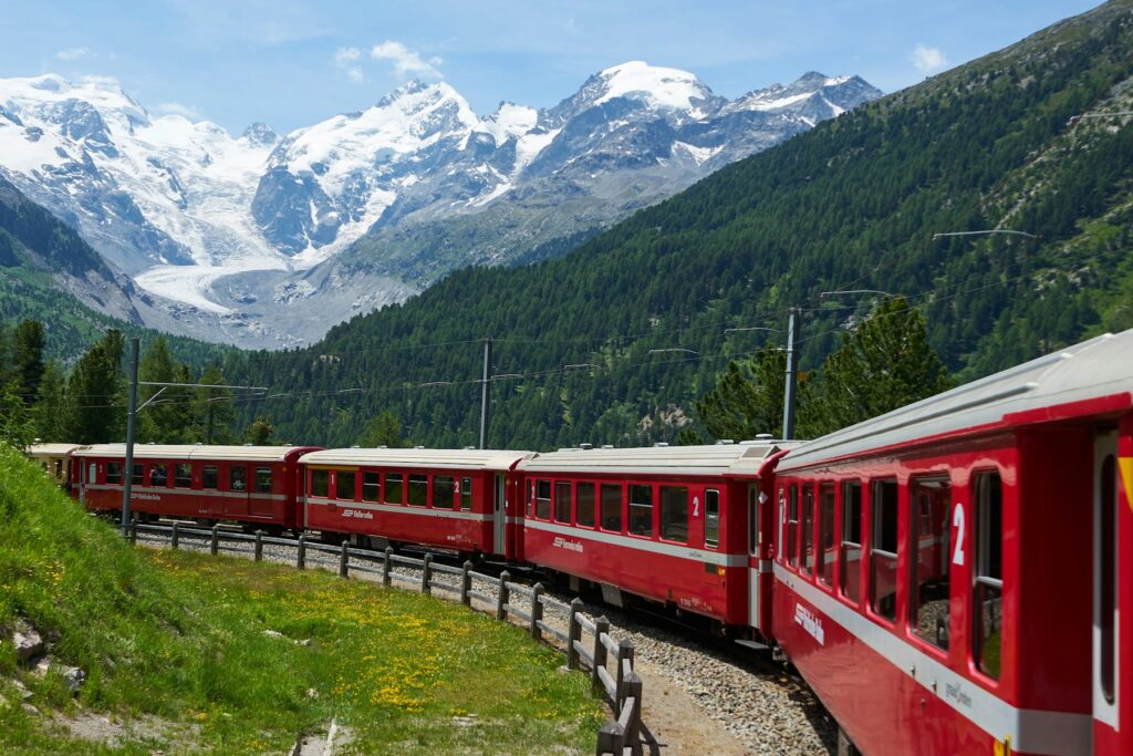 Trem vermelho fazendo uma curva num vale verde dos Alpes suíços, com montanhas nevadas e uma geleira ao fundo